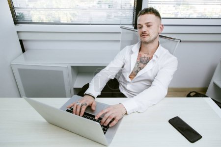 Téléchargez les photos : Cagliari, Italie - 20-03-2018 : Gérant blanc avec chemise blanche et tatouages sur tout le corps assis sur le bureau devant l'ordinateur dans son bureau - en image libre de droit