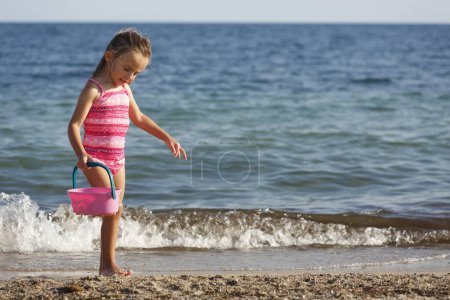 Téléchargez les photos : Petite fille blonde aux yeux verts joue avec le sable à la mer en maillot de bain - en image libre de droit