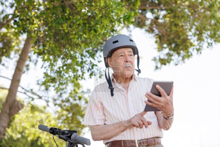 Photo for Elderly man with a protective helmet uses his tablet to surf the internet sitting next to his electric scooter in a park - Royalty Free Image