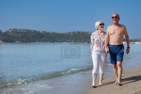 Photo for An elderly married couple affectionately hold hands as they walk along the shoreline of a beach - Royalty Free Image