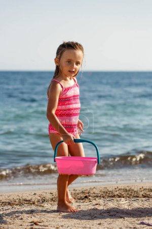 Foto de Niña rubia con ojos verdes juega con la arena en el mar en un traje de baño - Imagen libre de derechos
