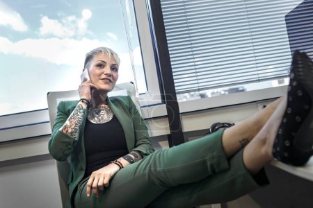 Téléchargez les photos : Gestionnaire blonde tatouée, vêtue d'une veste verte au bureau, parle sur son téléphone portable assis comme un patron dans sa chaise. - en image libre de droit