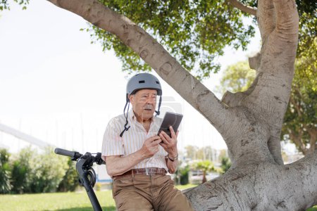 Téléchargez les photos : Homme âgé avec un casque de protection utilise sa tablette pour surfer sur Internet assis à côté de son scooter électrique dans un parc - en image libre de droit