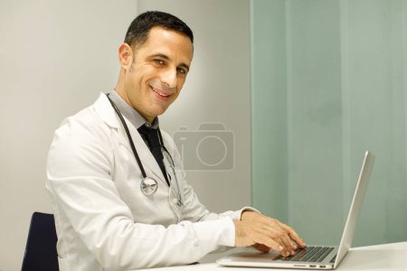 Photo for Dark-haired doctor with black hair in a white coat, working smiling at the computer in his surgery - Royalty Free Image