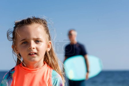 Foto de Pequeña rubia con ojos verdes juega con arena mientras su padre sostiene una tabla de surf en el fondo - Imagen libre de derechos