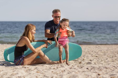Foto de Alegre familia que consta de padre, madre e hija se divierte jugando con una tabla de surf en la playa en el mar - Imagen libre de derechos