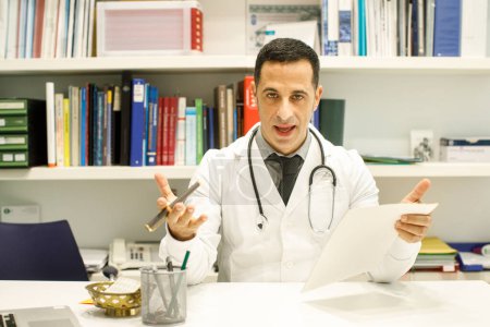 Photo for Black-haired doctor in a white coat reads data sitting in the desk of his medical office - Royalty Free Image