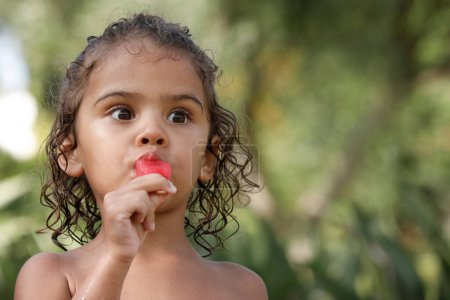 Photo for Cute portrait of a two year old girl of South American origin eating a fruit popsicle isolated on green nature background - Royalty Free Image
