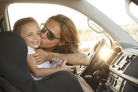 Téléchargez les photos : Femme au volant avec t-shirt blanc dans un contexte de vacances embrasse sa fille avec des expressions souriantes - en image libre de droit