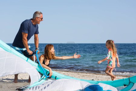 Foto de Familia compuesta por padre, madre y una hija prepara kitesurf en la playa listo para usarlo en el mar. - Imagen libre de derechos