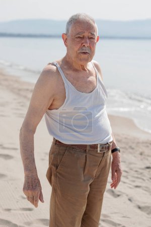Photo for An elderly gentleman dressed in a tank top by the sea is standing with a serious expression - Royalty Free Image