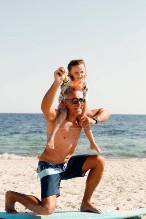 Téléchargez les photos : Papa avec des lunettes de soleil en maillot de bain enseigne les bases du surf à sa fille sur une plage balnéaire. - en image libre de droit