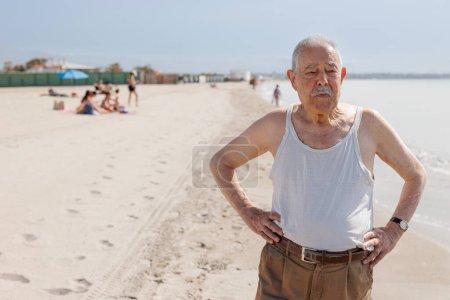 Foto de Un anciano caballero vestido con un tanque junto al mar está de pie con una expresión seria - Imagen libre de derechos