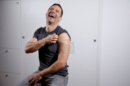 Photo for Dark-haired man with a shirt, sitting in a clinic, shows the arm with the patch after having the vaccine and gives a big smile - Royalty Free Image