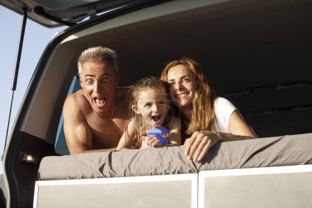 Téléchargez les photos : Famille heureuse composée de papa maman et fille, dans des vêtements de plage serrant joyeusement l'autre près du van au milieu d'une route - en image libre de droit