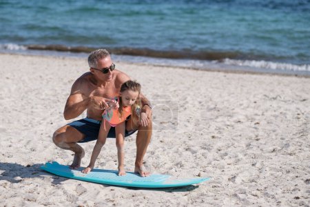 Téléchargez les photos : Papa avec des lunettes de soleil en maillot de bain enseigne les bases du surf à sa fille sur une plage balnéaire. - en image libre de droit