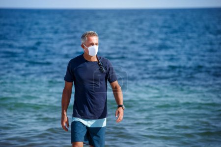 Foto de Hombre de 50 años con máscara protectora y camiseta azul se para en la playa y camina pacíficamente frente al mar - Imagen libre de derechos