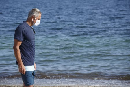 Foto de Hombre de 50 años con máscara protectora se para en la playa y parece desolado hacia el mar - Imagen libre de derechos