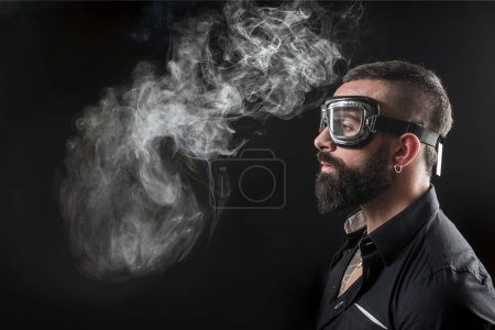 Téléchargez les photos : Portrait de fumeur bel homme barbu avec des tatouages sur fond sombre - en image libre de droit