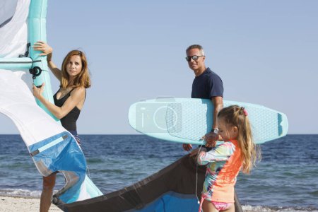 Foto de Familia compuesta por padre, madre y una hija prepara kitesurf en la playa listo para usarlo en el mar. - Imagen libre de derechos