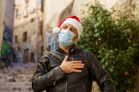 Photo for Man with Santa hat, leather jacket and surgical mask, puts his hand to his heart in an urban context with murals and graffiti on the walls - Royalty Free Image