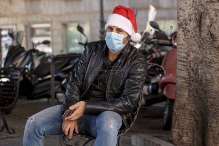 Téléchargez les photos : Homme avec chapeau de Père Noël, veste en cuir et masque chirurgical, en milieu urbain - en image libre de droit