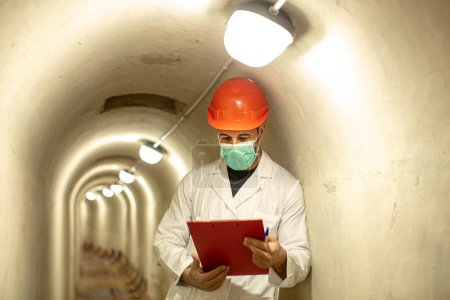Téléchargez les photos : Ingénieur avec casque de protection, masque chirurgical et manteau blanc tient un dossier rouge à l'intérieur d'un tunnel souterrain - en image libre de droit