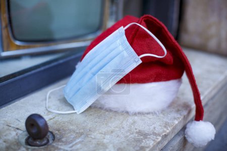 Téléchargez les photos : Chapeau de Père Noël avec un masque chirurgical à proximité et en arrière-plan derrière une vitrine il y a une télévision vintage - en image libre de droit