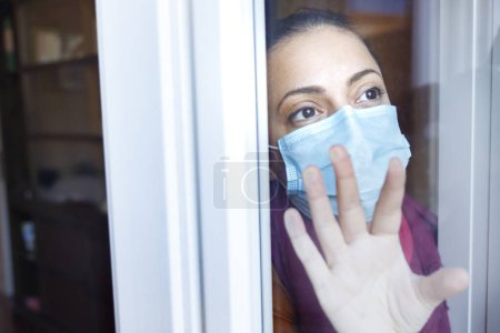 Photo for Young girl wears a protective mask inside the house and looks serious out the window - Royalty Free Image