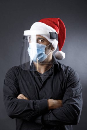 Téléchargez les photos : Homme avec chemise noire, masque chirurgical, bouclier protecteur en plexiglas et chapeau de Père Noël semble grave, isolé sur fond noir - en image libre de droit