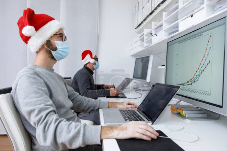 Foto de Hombres con máscaras médicas trabajando en la oficina con un ordenador portátil, concepto de Navidad, año nuevo, pandemia coronavirus - Imagen libre de derechos