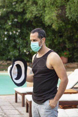 Téléchargez les photos : Homme aux cheveux bruns en tenue estivale avec chapeau et masque chirurgical se détend dans la piscine d'un hôtel - en image libre de droit