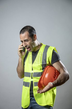 Téléchargez les photos : Travailleur avec casque et gilet jaune utilise son téléphone portable, isolé sur fond clair - en image libre de droit