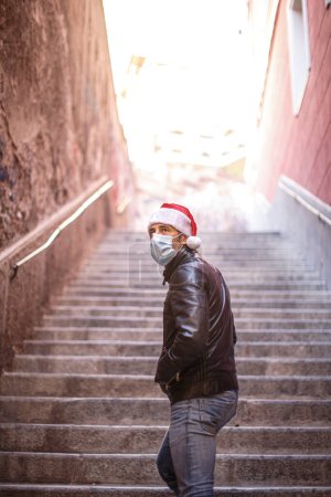 Téléchargez les photos : Homme au chapeau de Père Noël, veste en cuir et masque chirurgical, semble triste isolé sur un escalier - en image libre de droit