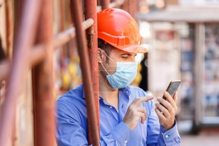 Photo for Engineer stands near a city construction site wearing a protective helmet and a surgical mask while using his cellphone - Royalty Free Image