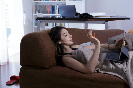 Téléchargez les photos : Belle fille brune regarde sa tablette tout en étant couché dans le canapé - en image libre de droit
