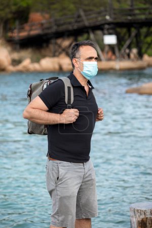 Foto de Hombre moreno en traje de verano y mochila camina por una ciudad balnearia con máscara protectora - Imagen libre de derechos