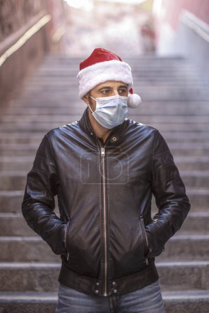 Photo for Man in Santa hat, leather jacket and surgical mask, looks sad isolated on a staircase - Royalty Free Image