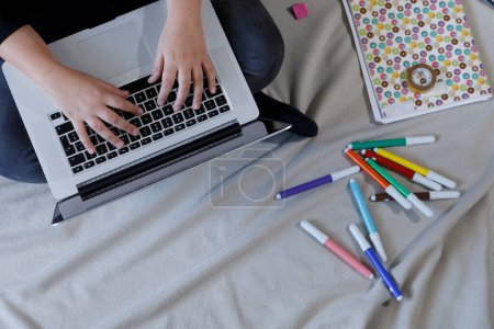 Téléchargez les photos : Mains détail de la jeune femme assise sur le lit écrivant sur l'ordinateur avec des stylos feutre à proximité et un carnet - en image libre de droit