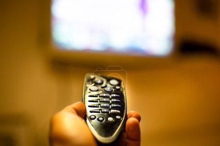 Téléchargez les photos : Télécommande au premier plan tenue par une main et un téléviseur flou en arrière-plan - en image libre de droit