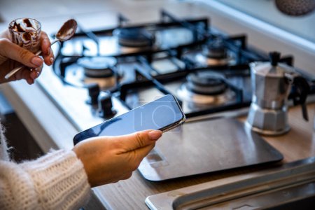 Foto de Mano de chica revisando un teléfono inteligente mientras en la cocina preparando café en la moka olla en el fondo - Imagen libre de derechos