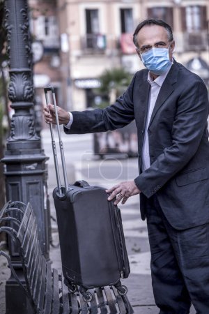 Photo for Tourist dressed in suit with protective face mask and trolley suitcase is in urban context - Royalty Free Image