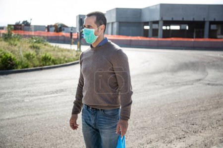Téléchargez les photos : Casual habillé homme avec masque protecteur et sac à provisions traverse la rue - en image libre de droit