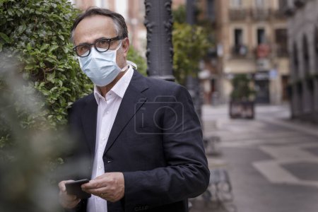 Téléchargez les photos : Portrait d'un homme d'âge moyen vêtu d'une veste avec masque protecteur, debout sur un trottoir avec de la verdure publique derrière - en image libre de droit