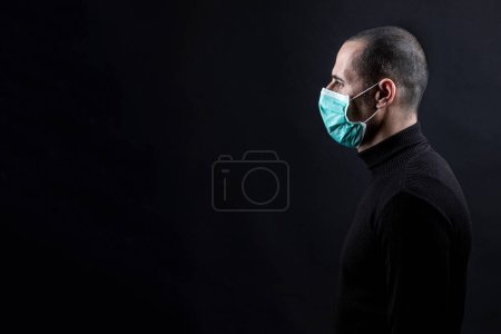 Photo for Man with shaved hair wearing a surgical mask, black sweater, is photographed in profile, isolated on a black background - Royalty Free Image