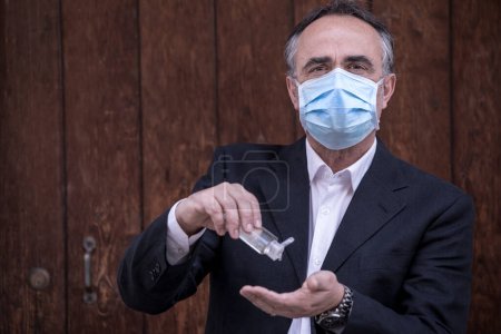 Photo for Man dressed in suit with protective face mask uses a hand sanitizer - isolated on wooden background - Royalty Free Image