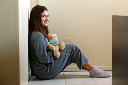 Photo for Girl in pajamas sitting at home holds a teddy bear in her hand wearing the protective mask against coronavirus - Royalty Free Image