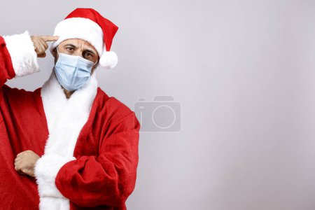 Téléchargez les photos : Père Noël avec masque chirurgical pointe son doigt vers sa tête dans la folie, isolé sur fond blanc - en image libre de droit