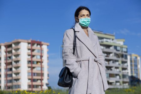 Foto de Chica morena con abrigo y máscara facial, en los edificios de fondo de un barrio de la ciudad y el cielo azul - Imagen libre de derechos
