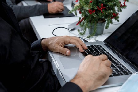 Foto de Mano de hombre sosteniendo una tarjeta de crédito azul sentada frente a la computadora en el contexto navideño y con una máscara quirúrgica a su lado - Imagen libre de derechos